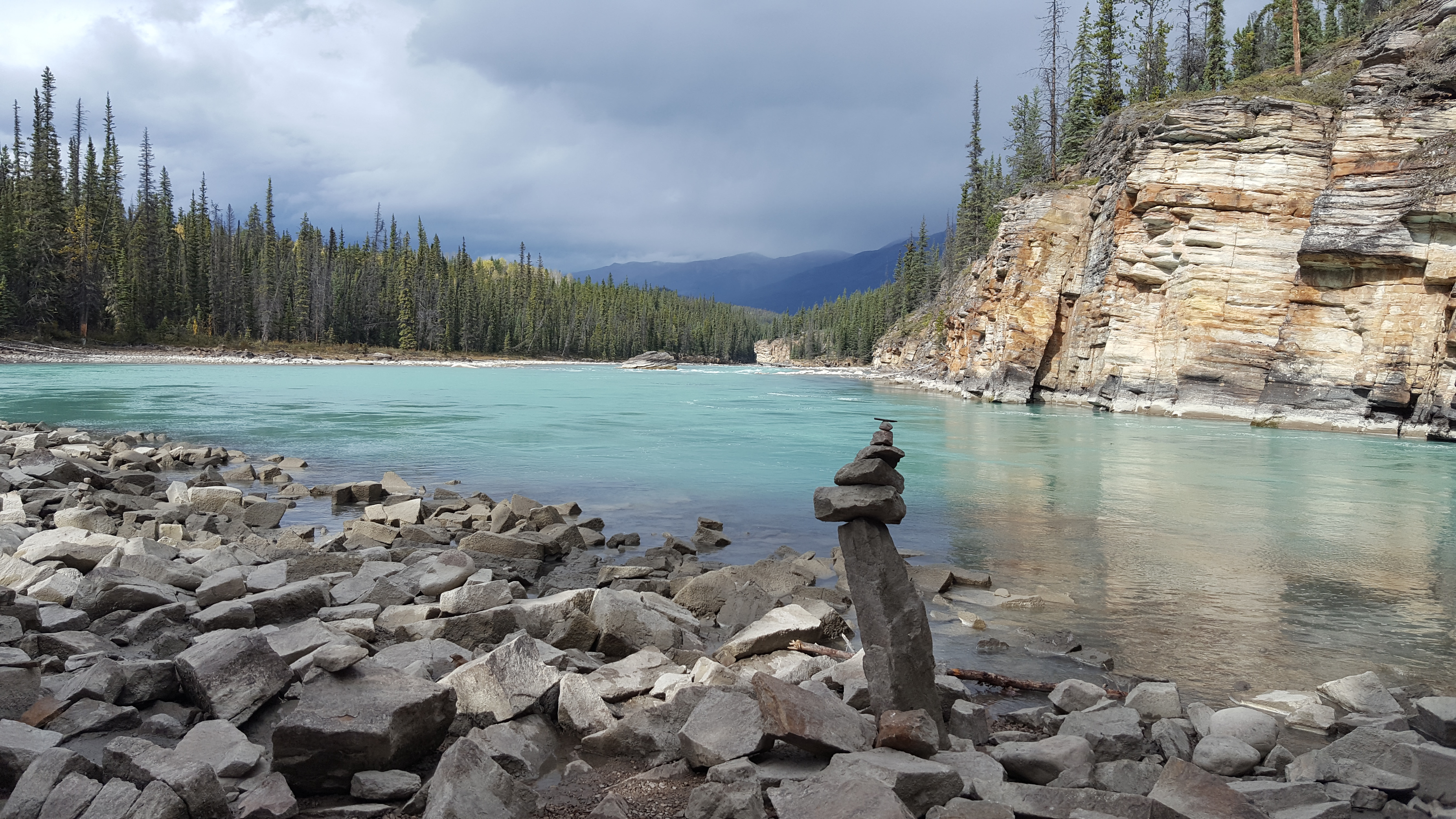 Image of clear blue lake with pine trees on the opposite bank and stones piled at the foreground